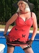 Blonde mature hottie in stockings, red dress and hat poses near swimming pool